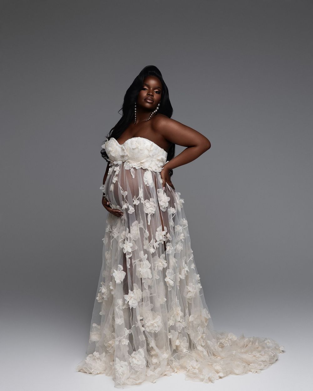 Pregnant woman in elegant white floral tulle maternity gown posing gracefully in a professional studio photo shoot.
