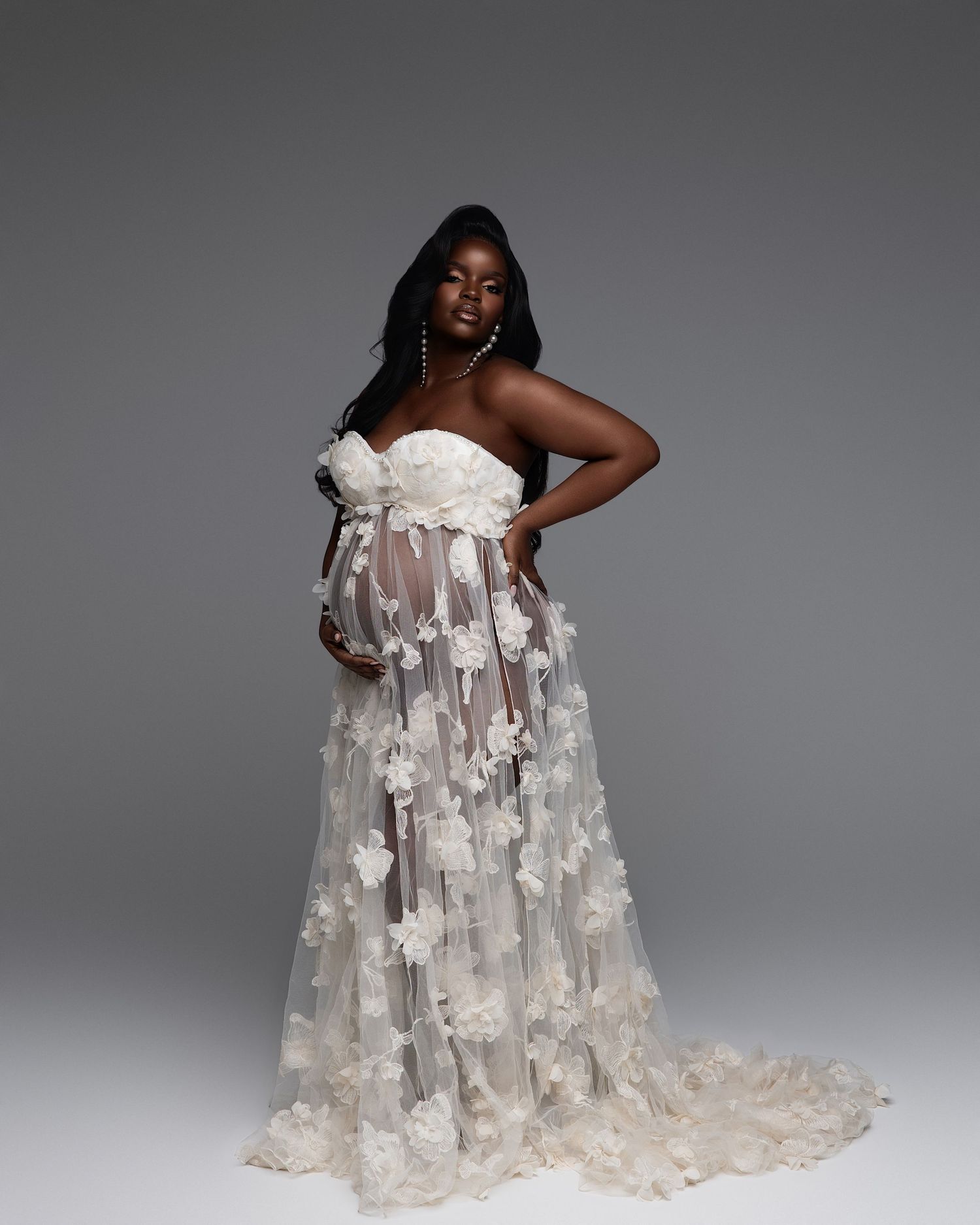 Pregnant woman in elegant white floral tulle maternity gown posing gracefully in a professional studio photo shoot.