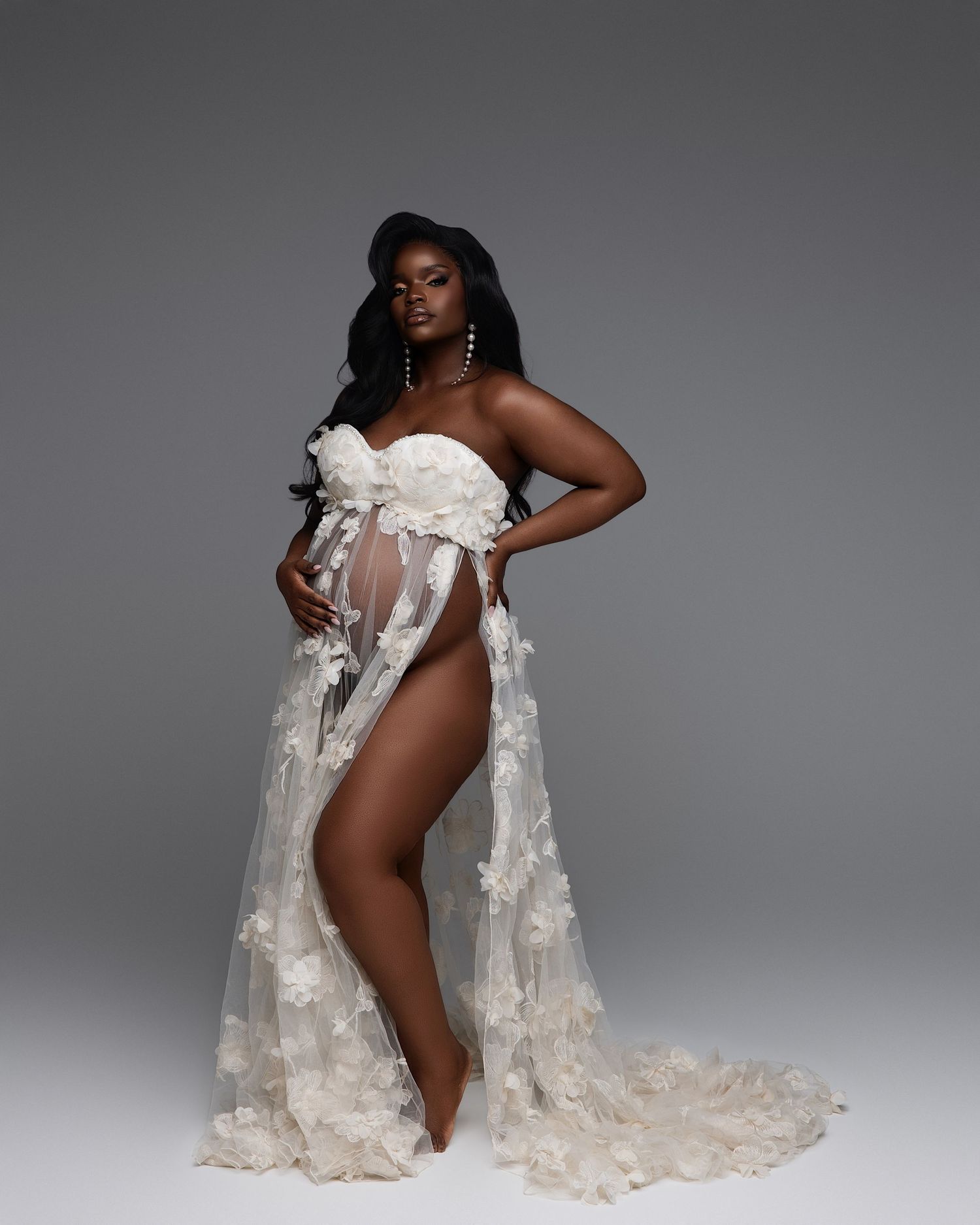 Pregnant woman in white floral sheer maternity gown poses gracefully against gray studio background.