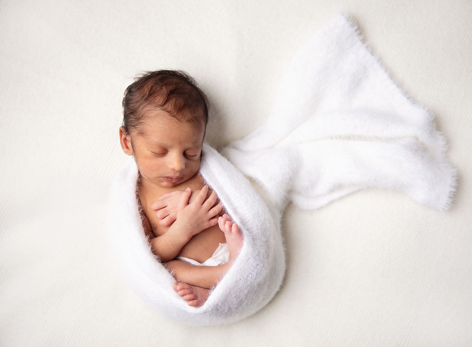 Newborn baby peacefully sleeping wrapped in a soft white blanket on a white background. During a newborn photoshoot in Bromley