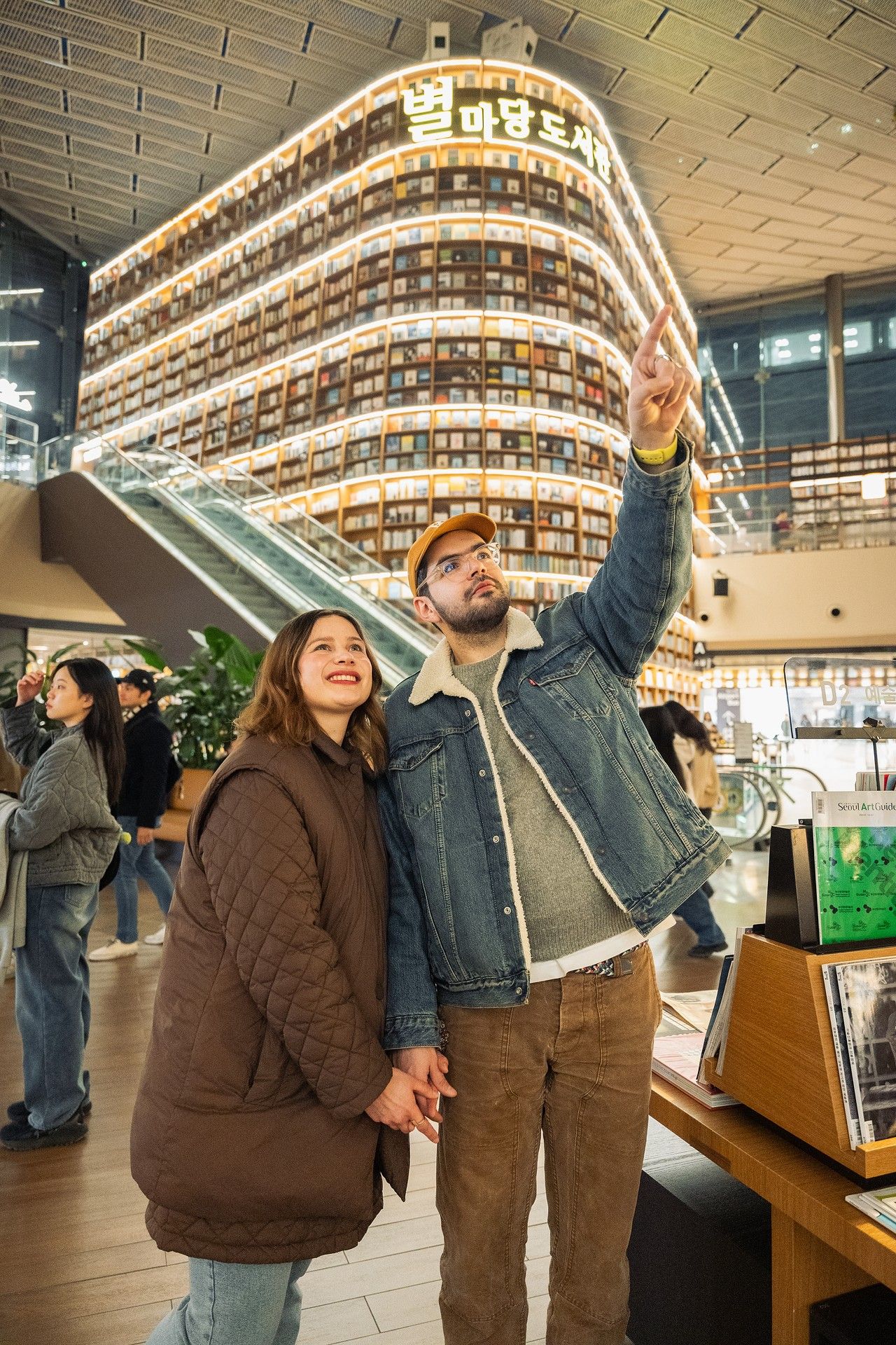 Couple holding hands at Starfield Library in COEX Mall, Seoul, man pointing upward at illuminated bookshelves.