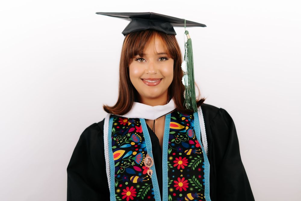 Smiling graduate in black cap and gown with colorful floral stole and green tassel on white background.