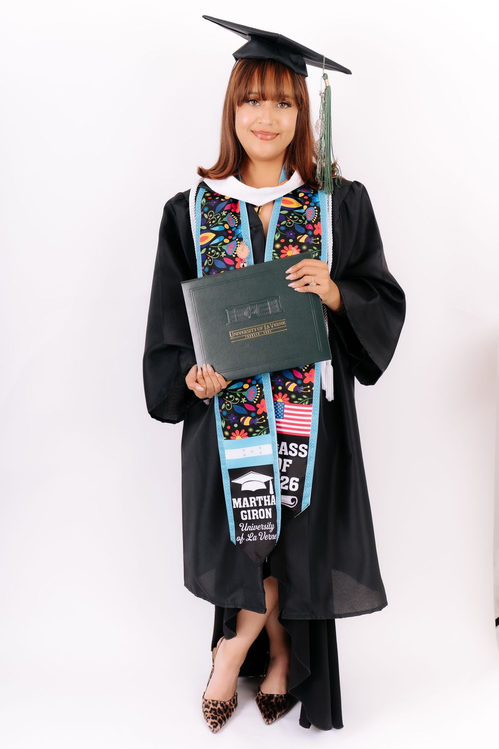 Young woman in black graduation gown and cap holds diploma, wearing colorful stole with name Martha Giron, Class of 2026.