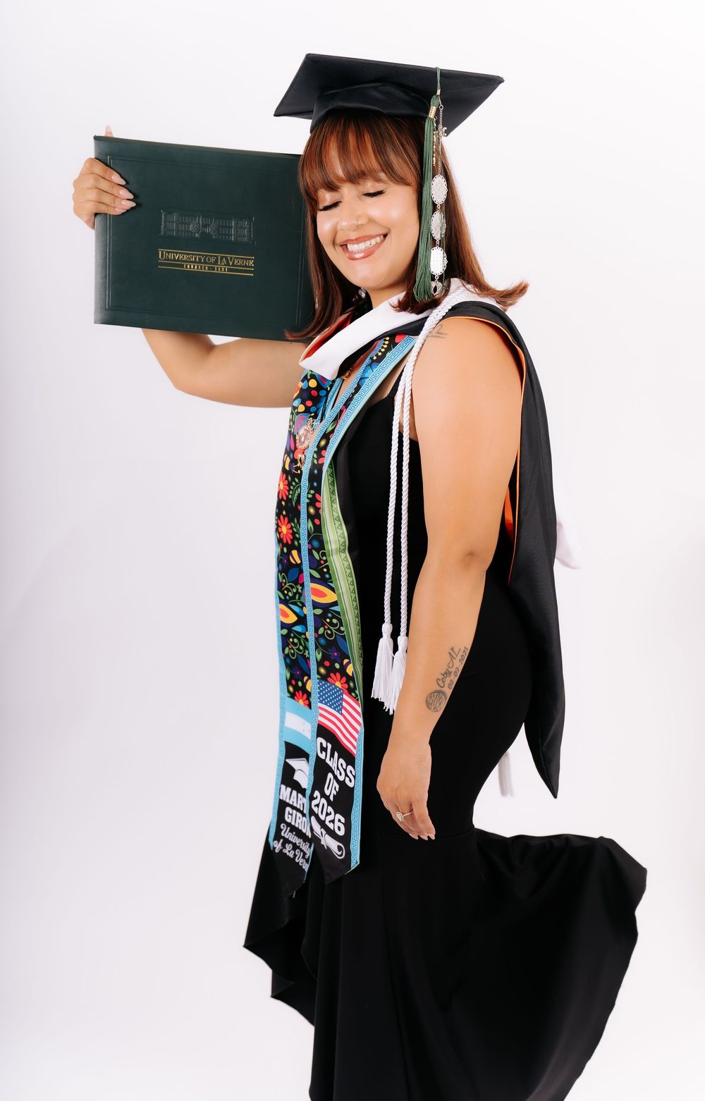 Smiling graduate in cap and gown holding diploma, wearing colorful stole and Class of 2025 sash, white background.