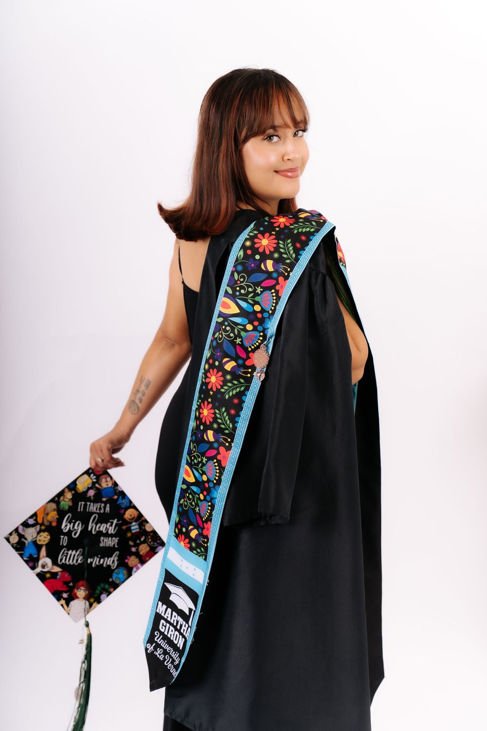 Graduate in black gown and floral sash holds decorated cap reading 'It takes a big heart to shape little minds'.