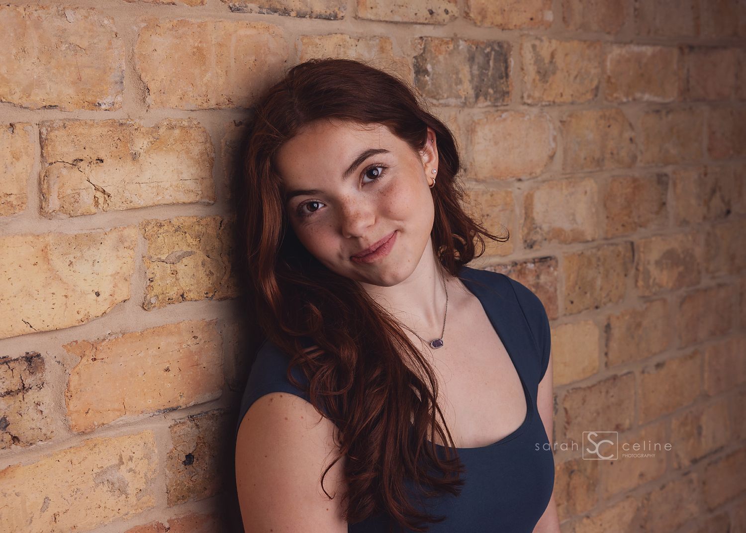 Young woman with long red hair smiling against a rustic brick wall in a blue top.