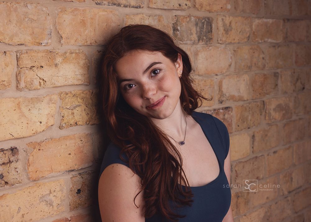 Young woman with long red hair smiling against a rustic brick wall in a blue top.