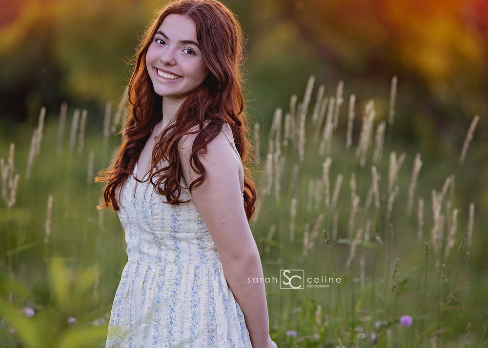 Young woman with red hair in white floral dress smiling in a sunlit meadow with tall grasses and wildflowers.