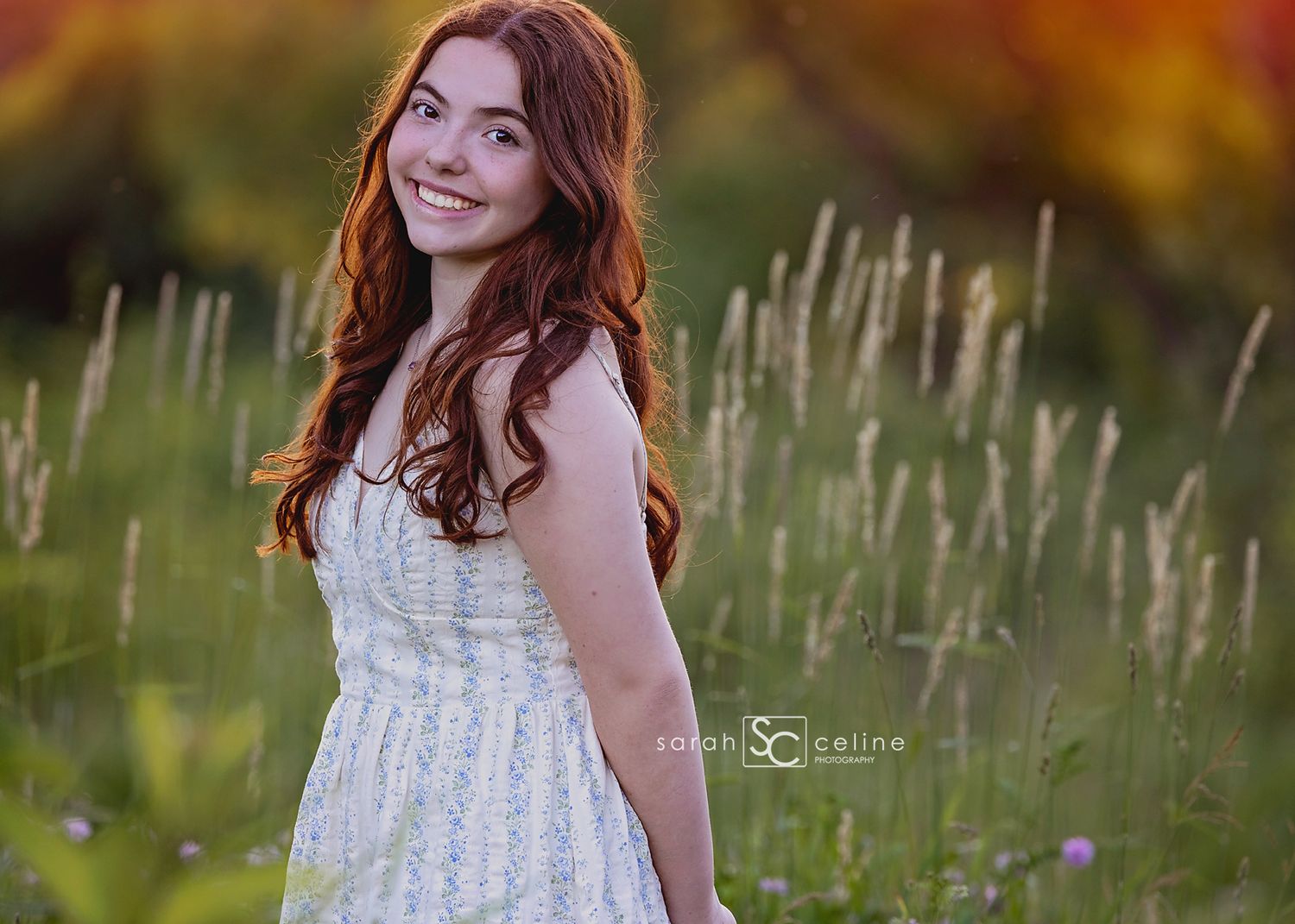 Young woman with red hair in white floral dress smiling in a sunlit meadow with tall grasses and wildflowers.