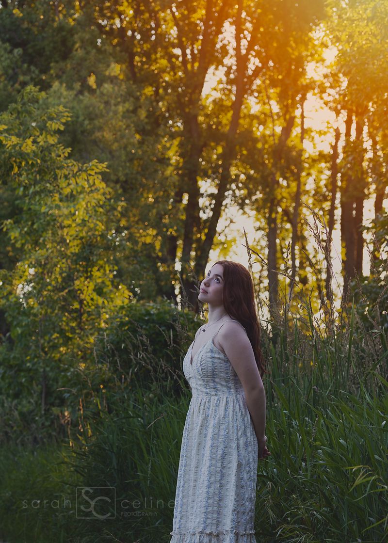 A young woman in a white floral dress stands in a sunlit forest clearing at golden hour, gazing upward.