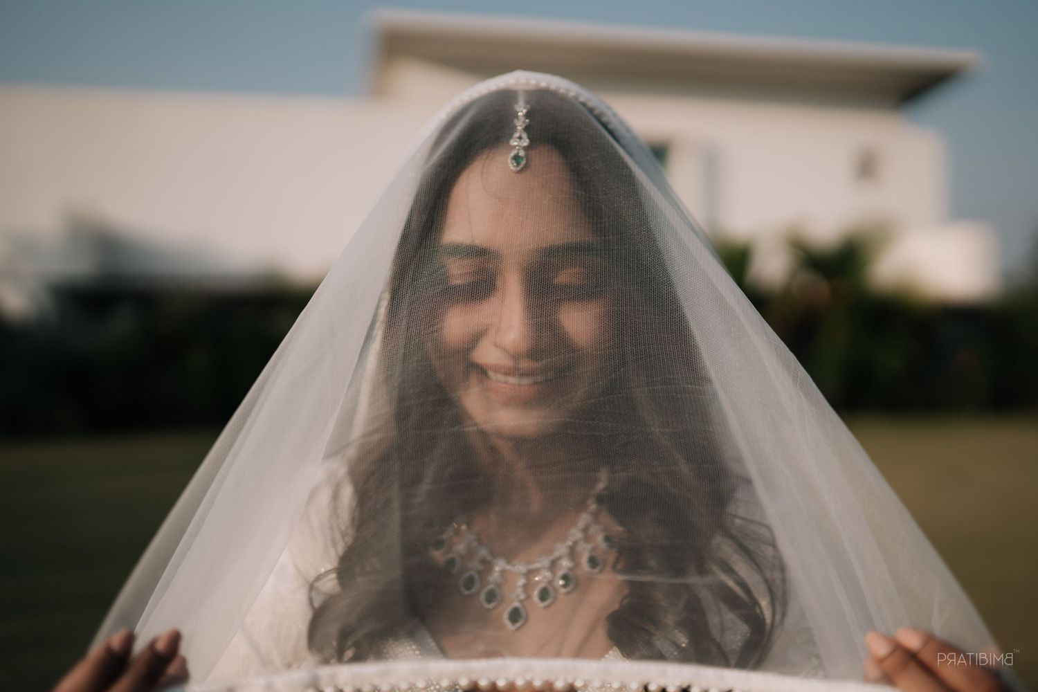 Indian bride smiling under a sheer white veil, wearing elegant jewelry and maang tikka at an outdoor wedding.