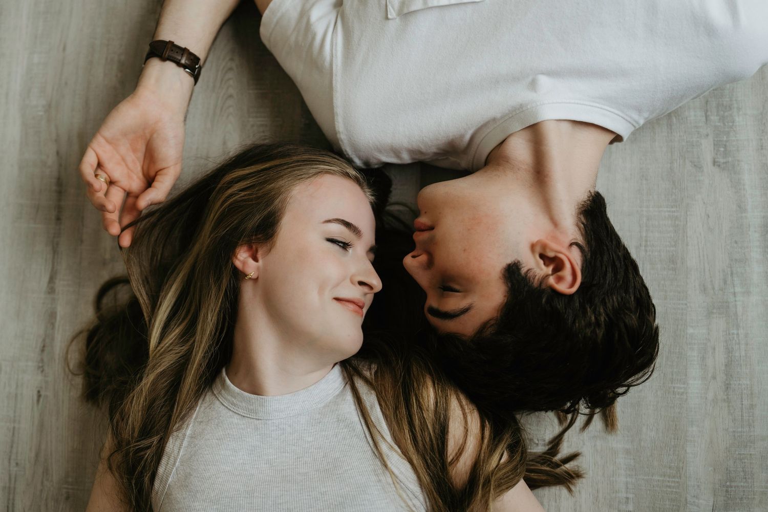 A young couple lying on the floor holding hands, smiling and gazing at each other in a romantic moment.