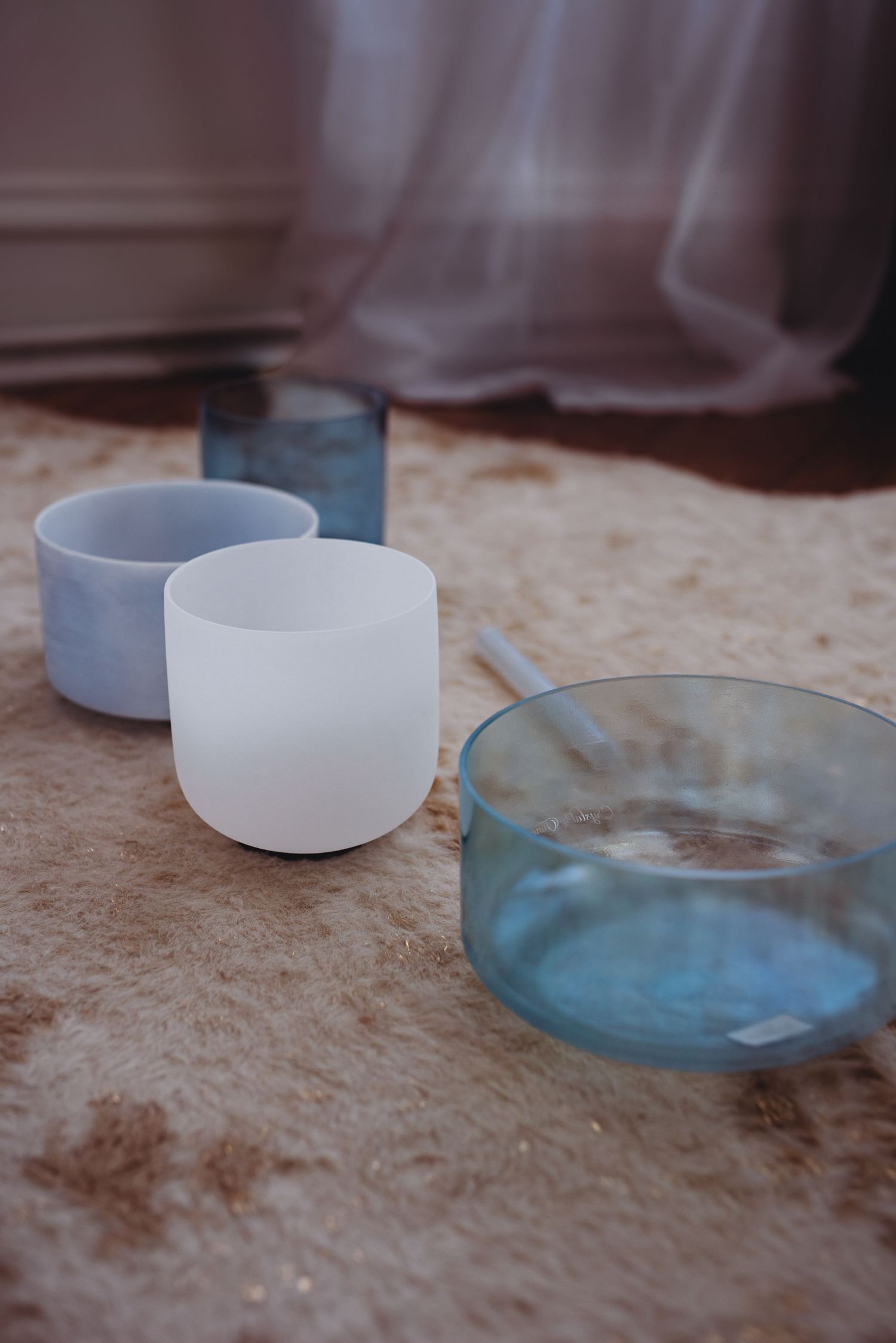 Person sitting on floor with crystal singing bowls and a blue glass, holding a mallet for sound healing.