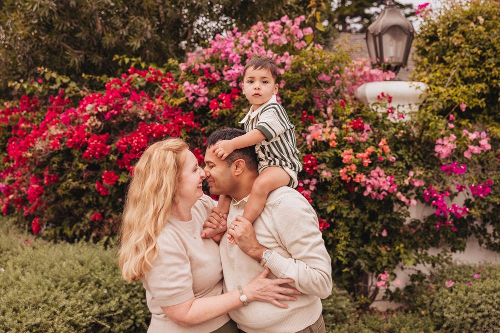 Family of three posing outdoors with vibrant pink bougainvillea flowers, child sitting on father's shoulders.
