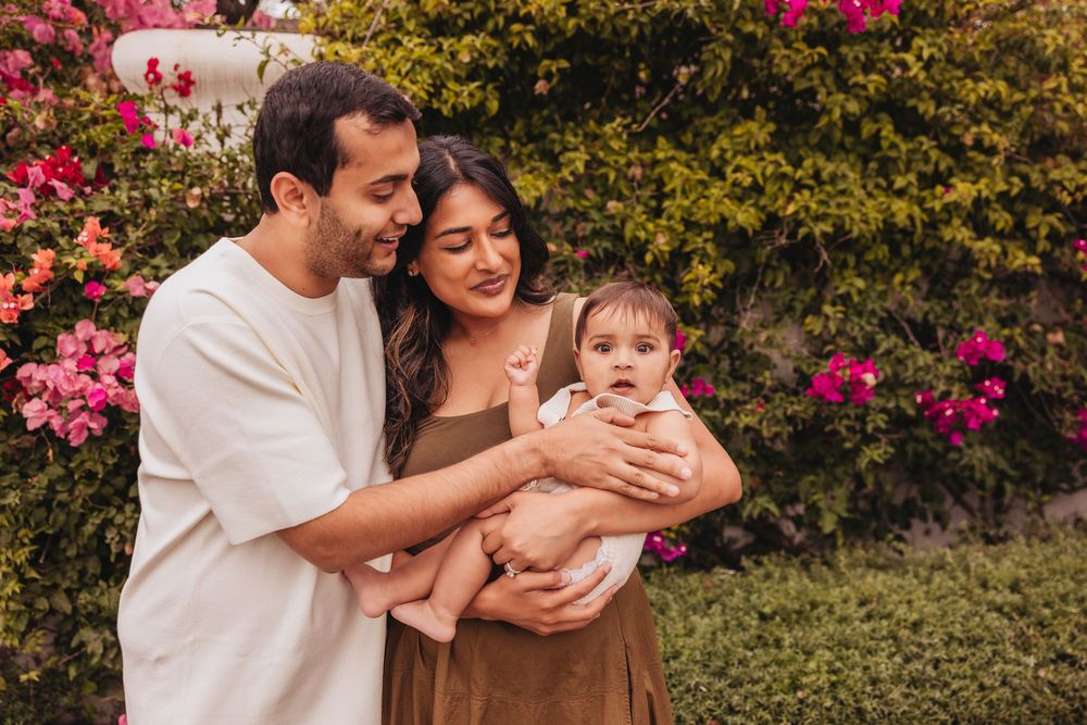 A young couple smiles while holding their baby outdoors, surrounded by vibrant pink flowering bushes.