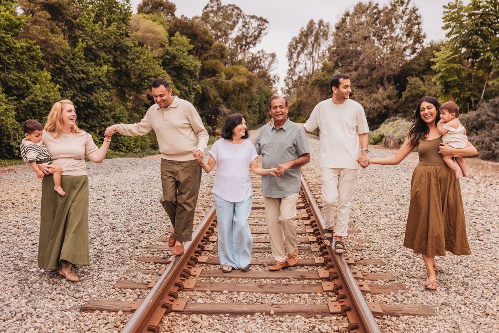 Family of seven holding hands on railroad tracks surrounded by lush green trees in a warm outdoor photo session.