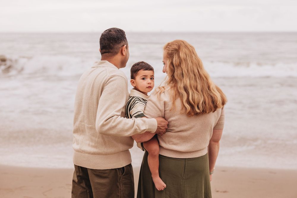 Family of three standing on a sandy beach, parents holding young child, facing the ocean waves.