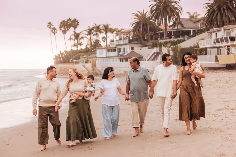 Multi-generational family walking barefoot on a sandy beach at sunset with palm trees and coastal homes in background.