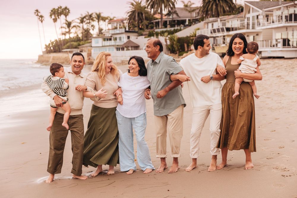 A joyful multigenerational family poses together on a sandy beach at sunset near a coastal home.