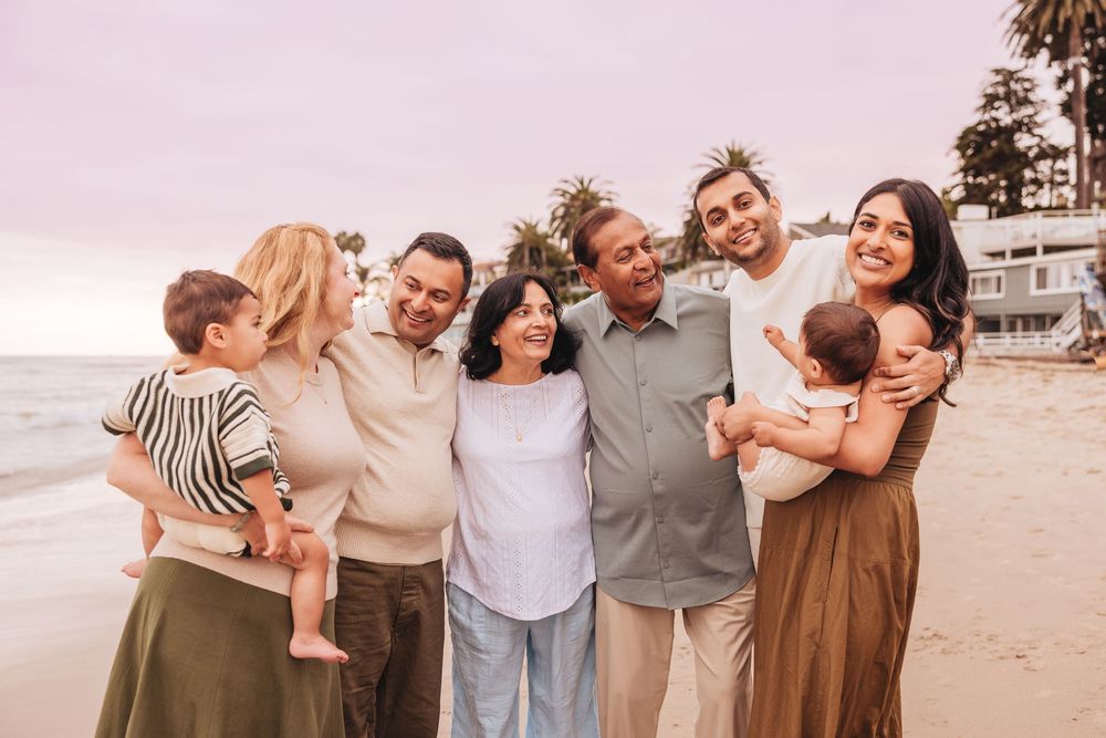 Multi-generational family smiling together on a beach at sunset, including grandparents, parents, and young children.