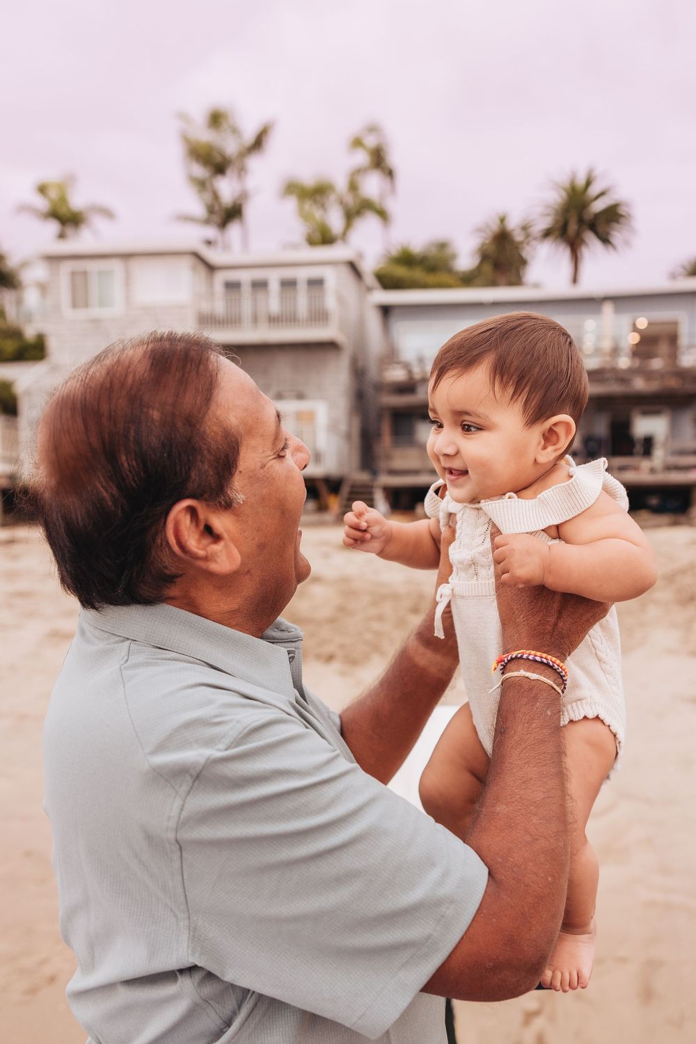 Grandfather joyfully lifts smiling baby on a sandy beach with palm trees and houses in the background.