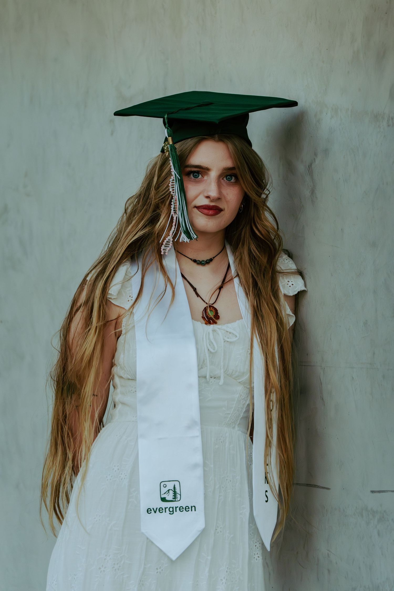 Graduate with long blonde hair poses in green mortarboard and white Evergreen stole indoors.
