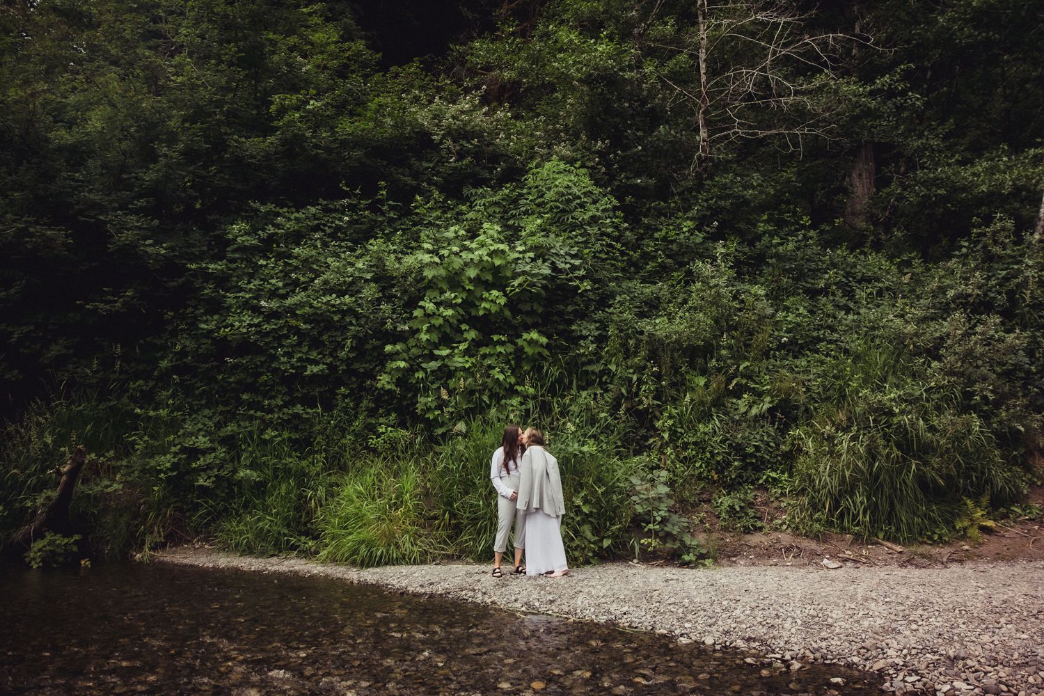 Couple in wedding attire standing on a rocky riverbank surrounded by lush green forest trees.