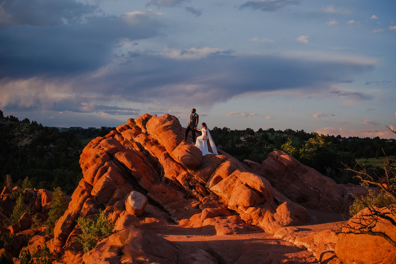 Couple in wedding attire standing atop red rock formations at sunset with dramatic cloudy sky in Colorado.