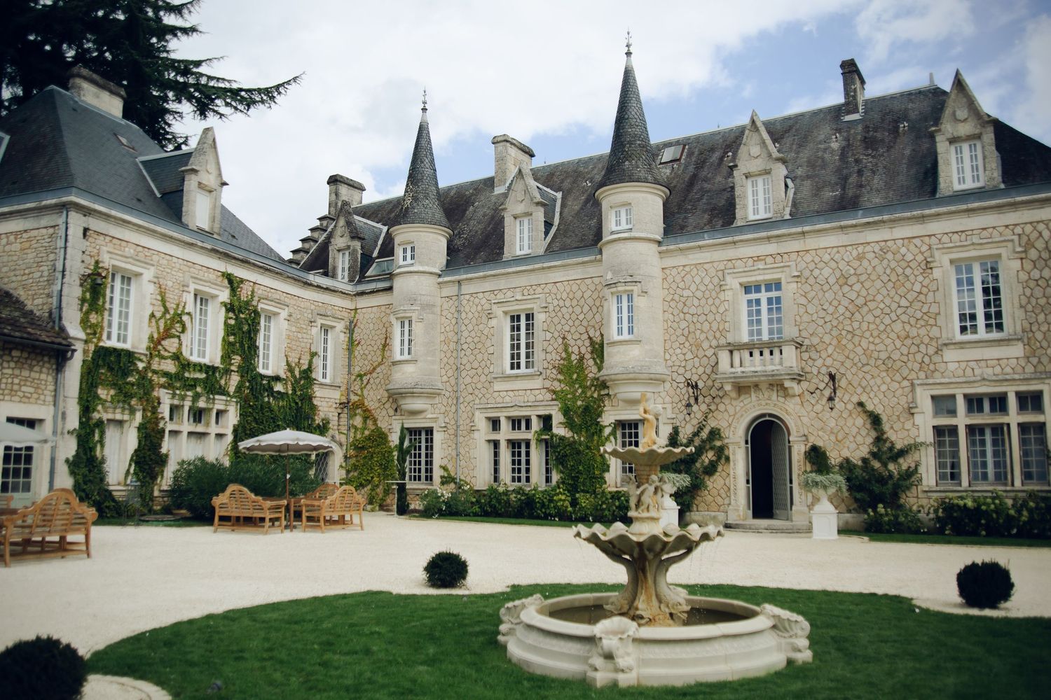 Elegant French château with stone façade, turrets, courtyard fountain, and manicured lawn on a sunny day.