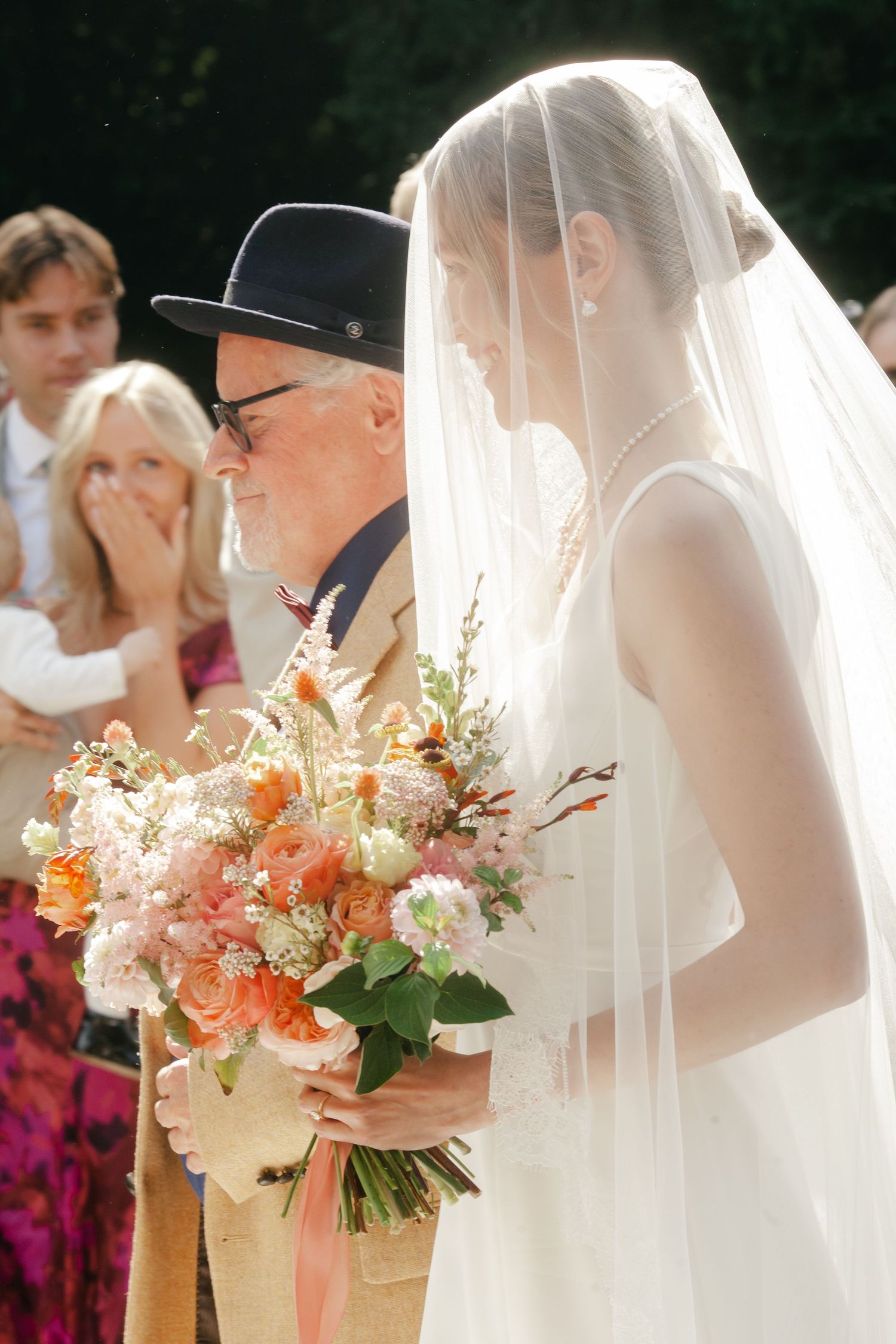 Bride in veil holding colorful bouquet with father during outdoor wedding ceremony.
