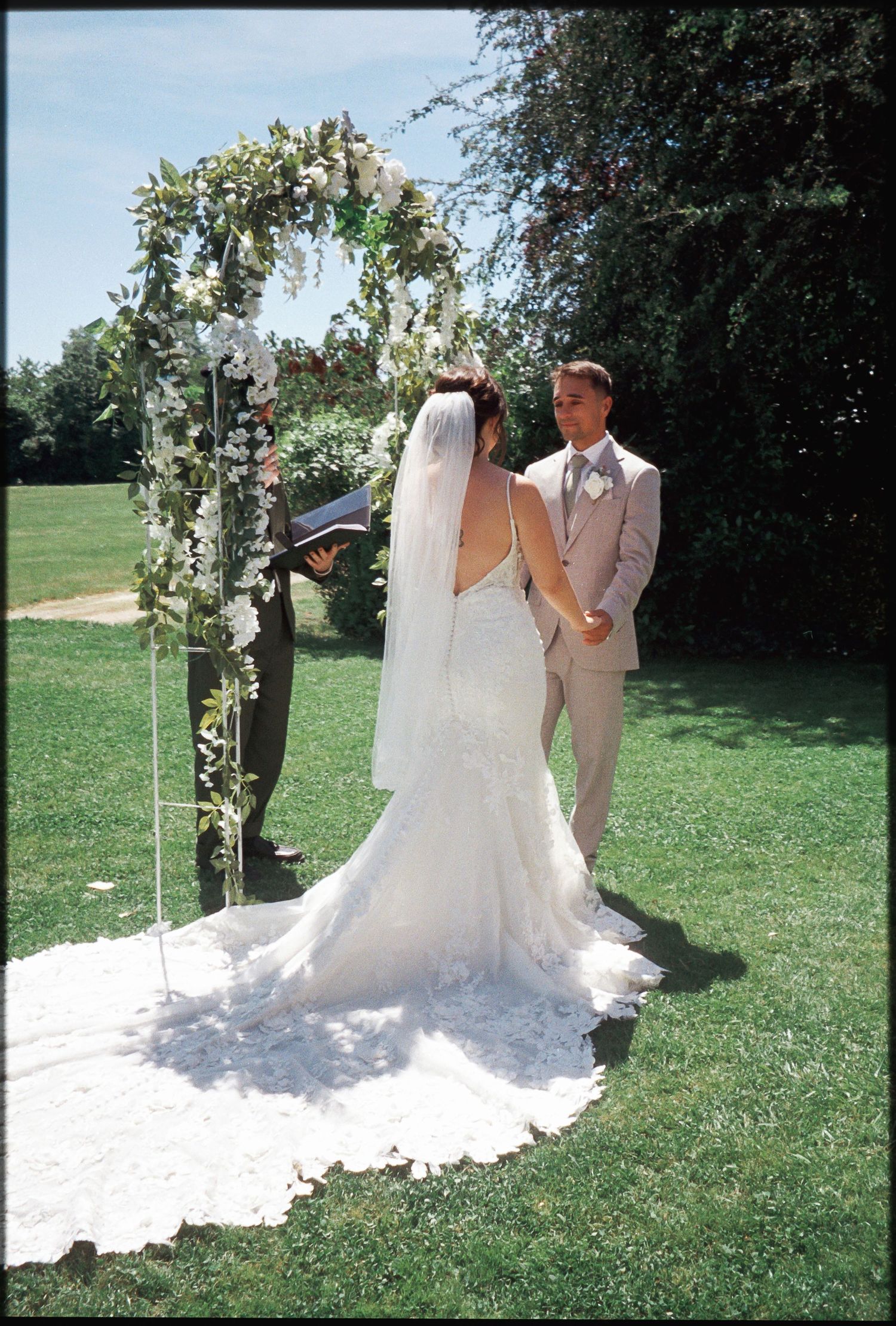 Outdoor wedding ceremony with bride in lace gown and groom in tan suit under floral arch on green lawn.