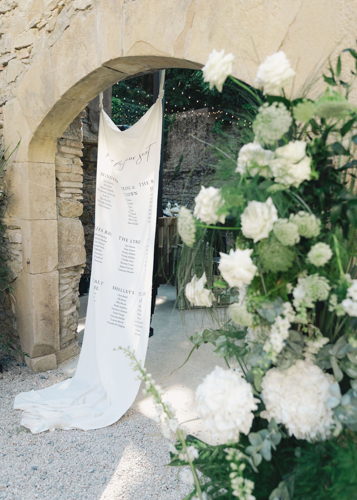 Elegant wedding seating chart banner hung in stone archway with lush white floral arrangements at French venue.