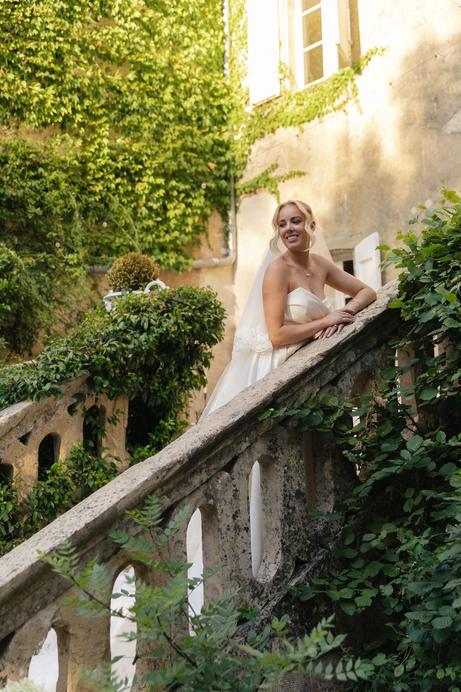 Smiling bride in white gown leans on stone balustrade of ivy-covered villa, posing elegantly outdoors.