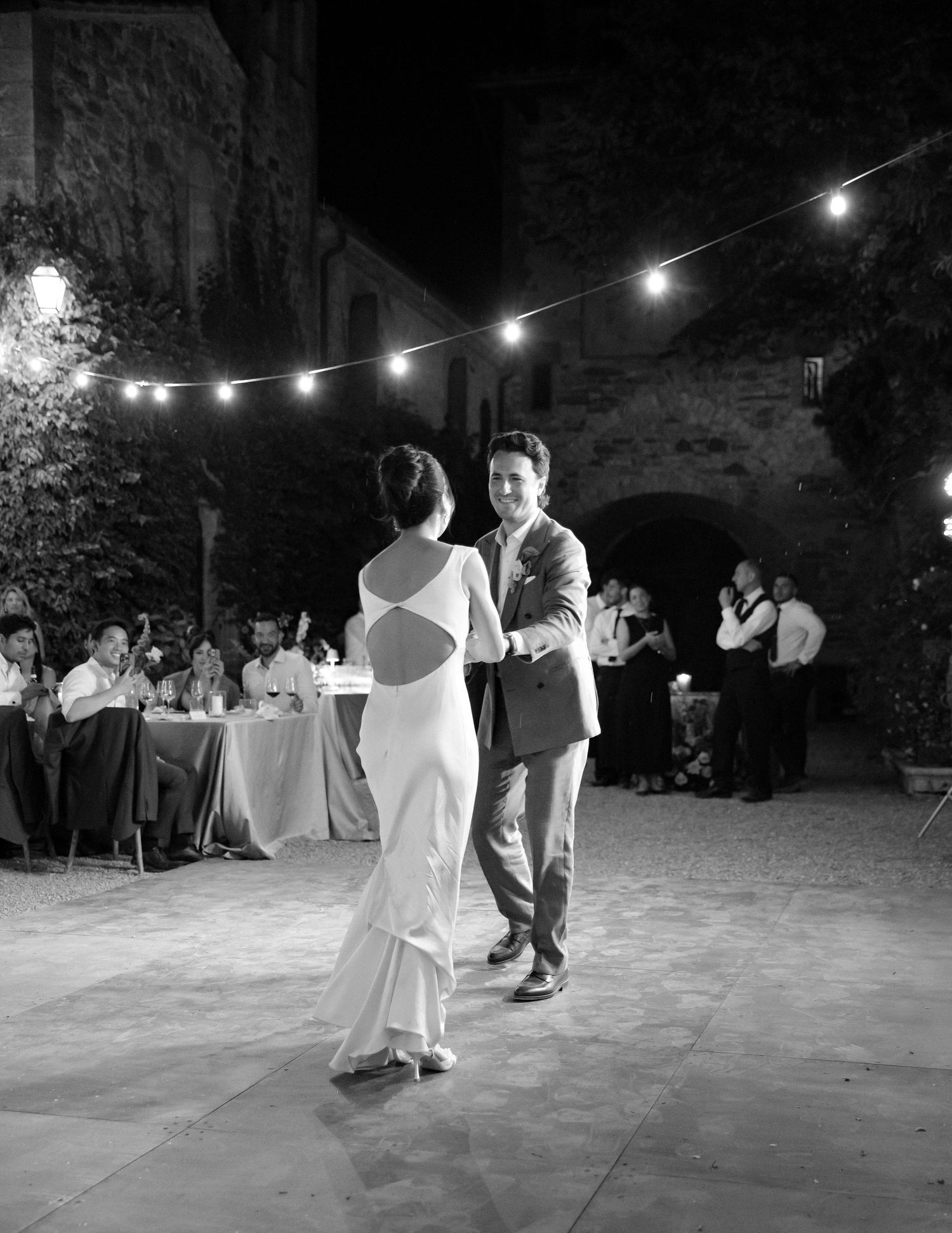 Couple shares first dance at outdoor wedding reception under string lights in elegant black and white photo.