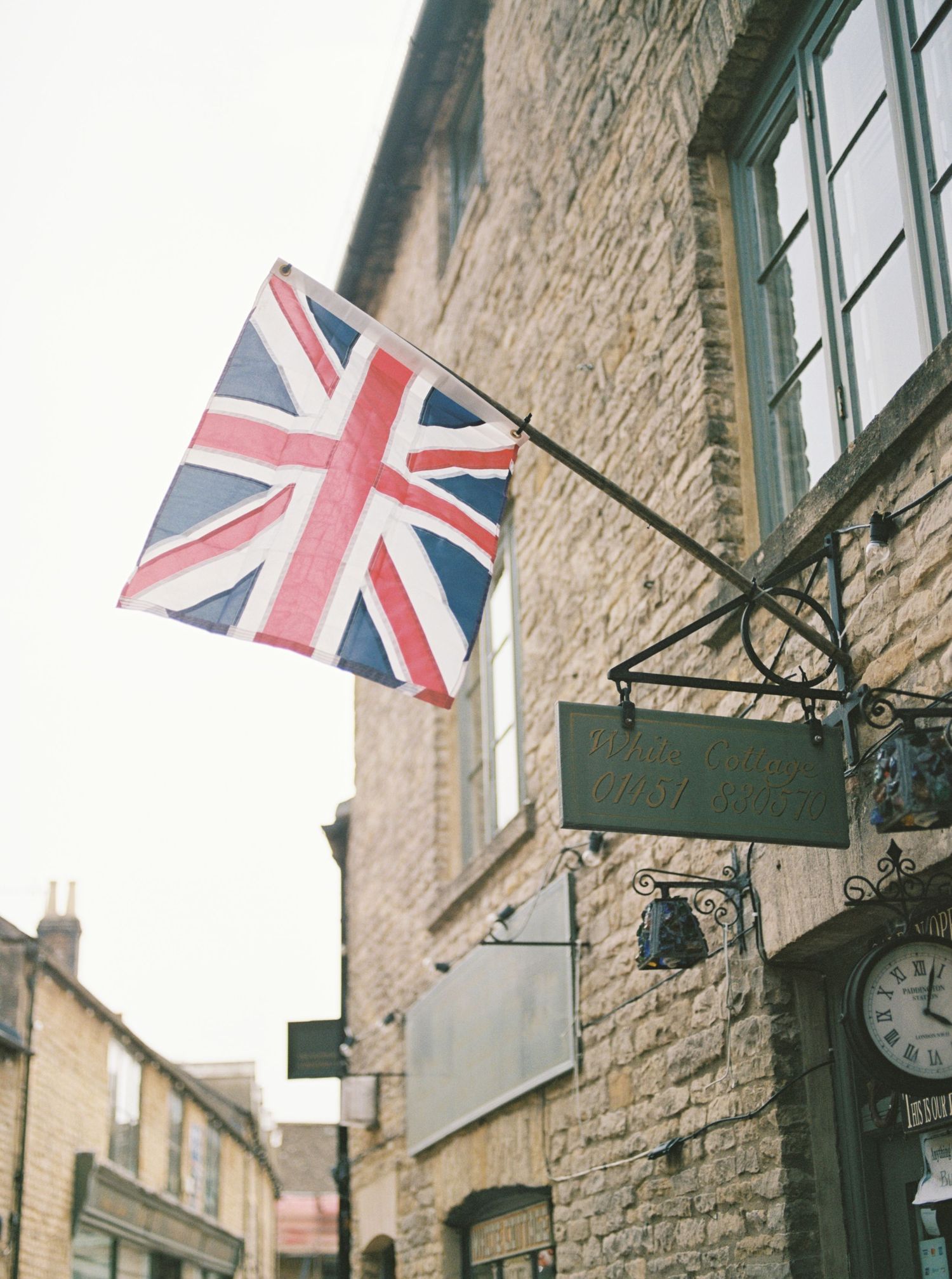 Union Jack flag flying outside a historic stone building with vintage shop signs and clock on a cobblestone street.