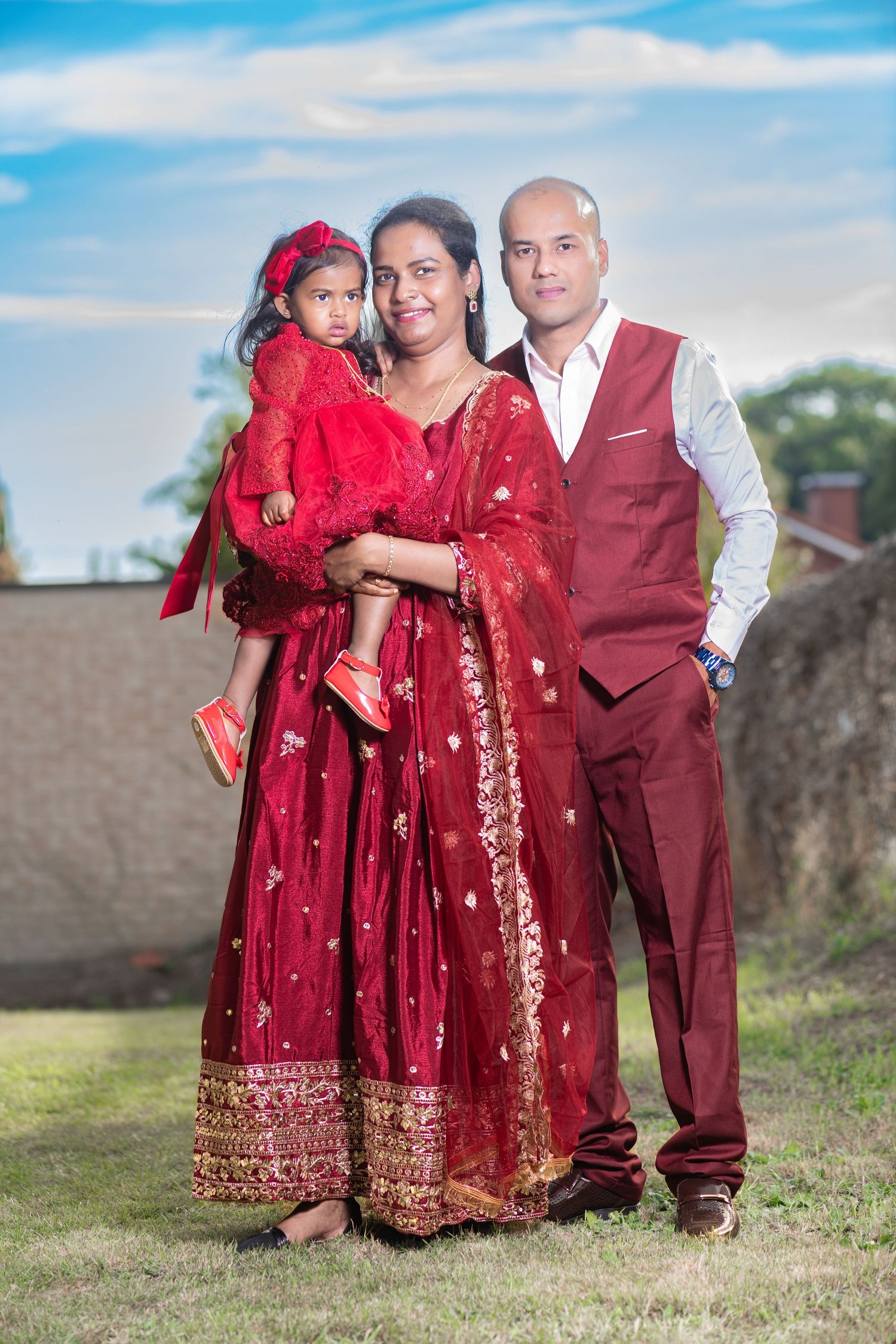 Indian family in matching red traditional attire posing outdoors, mother holding daughter, father standing beside them.