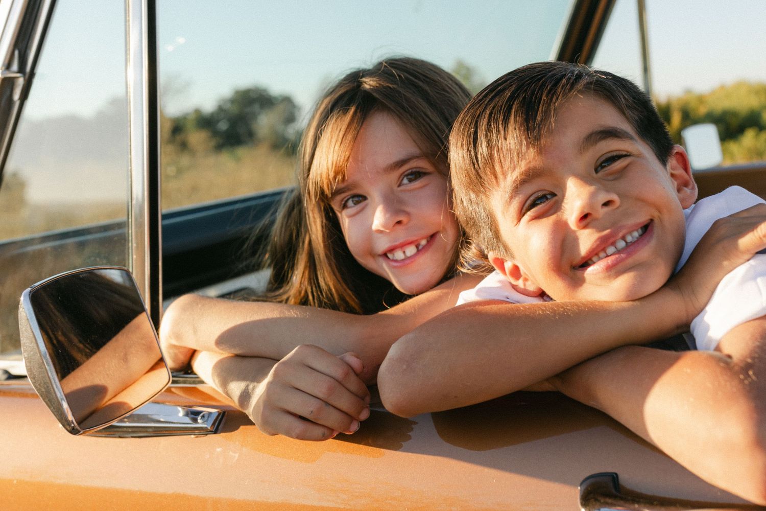Two happy children smiling and leaning out of a vintage orange car window on a sunny day.