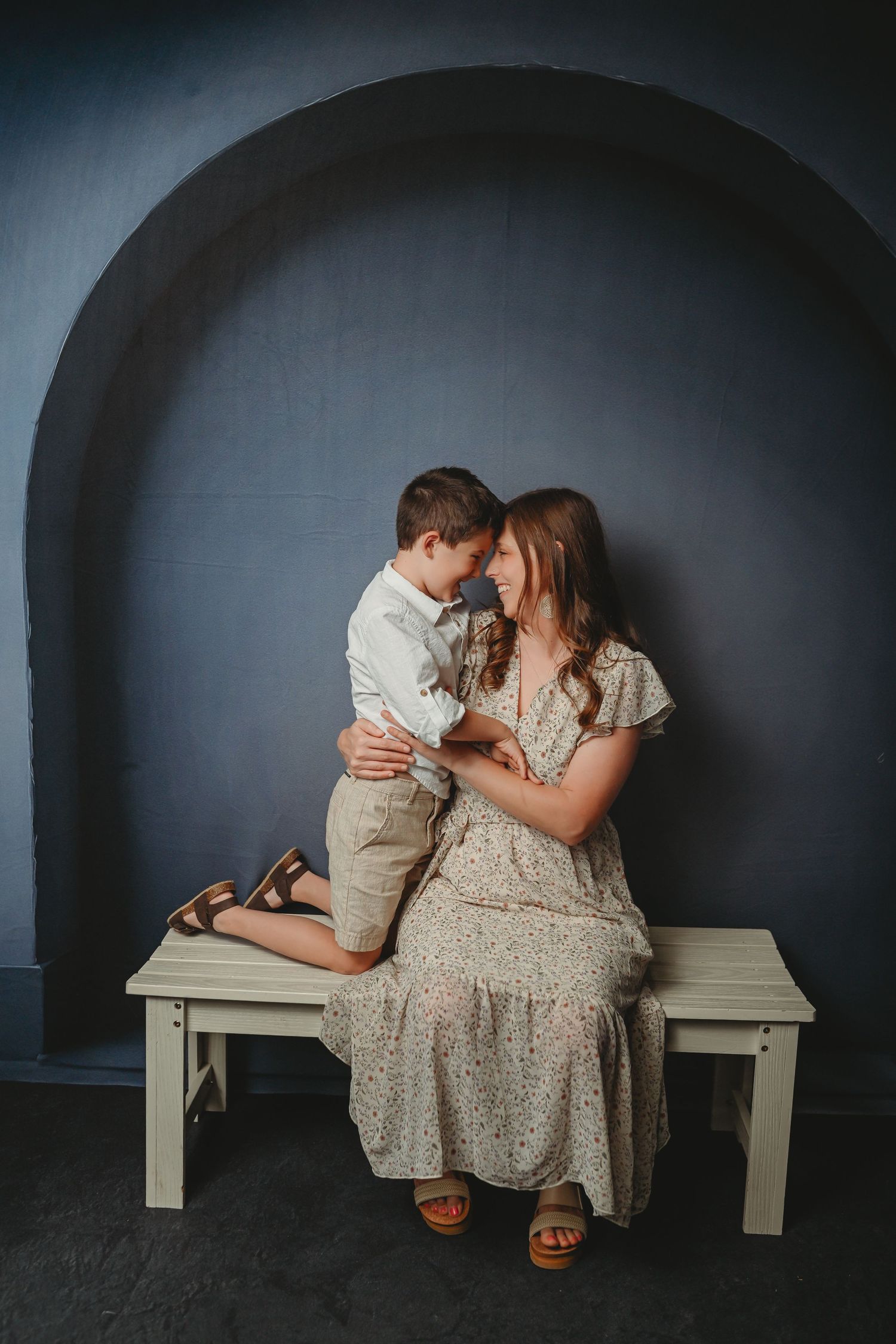 A mother in a floral dress sits on a bench hugging her young son in a studio with a gray arched wall backdrop.