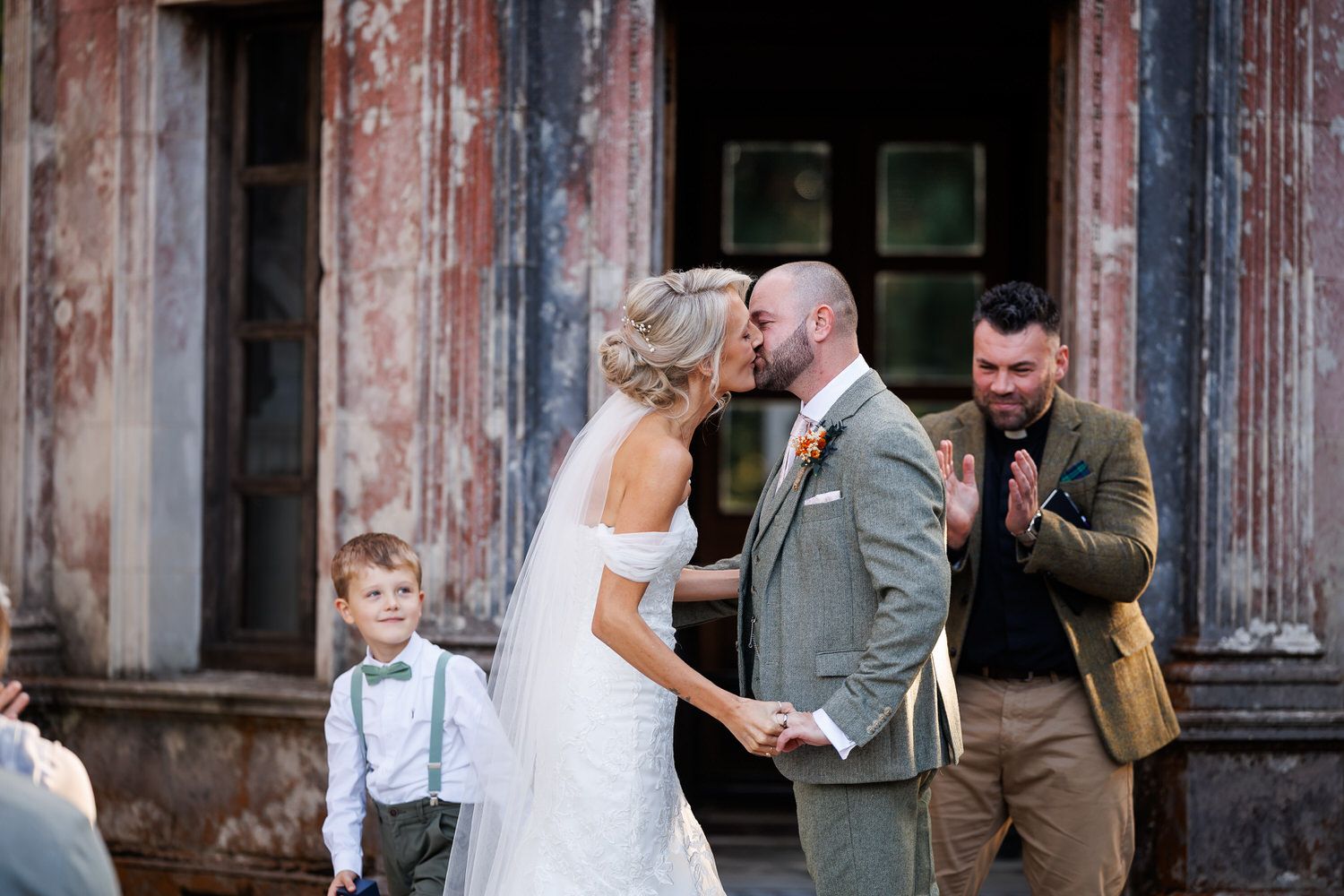 Bride and groom share first kiss at outdoor wedding ceremony while ring bearer and guest look on.