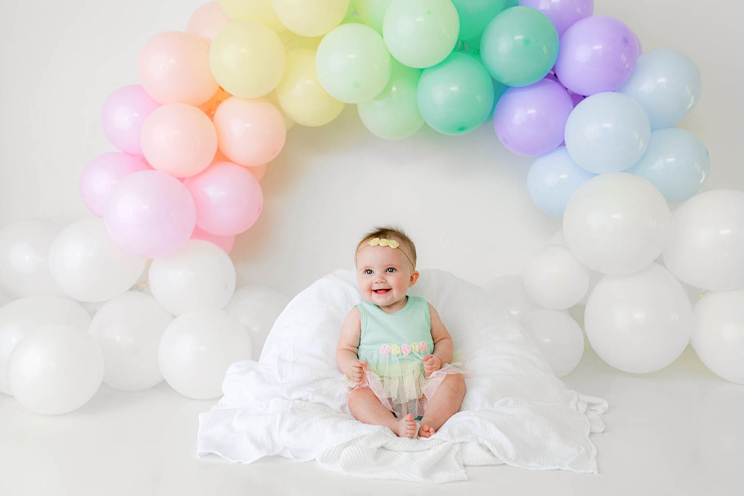 Smiling baby in mint outfit sitting under a pastel rainbow balloon arch on white backdrop.