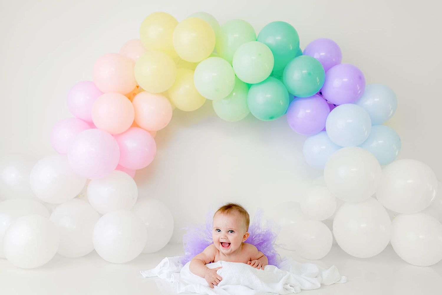 Happy baby in purple tutu lying under a colorful pastel rainbow balloon arch on white background.
