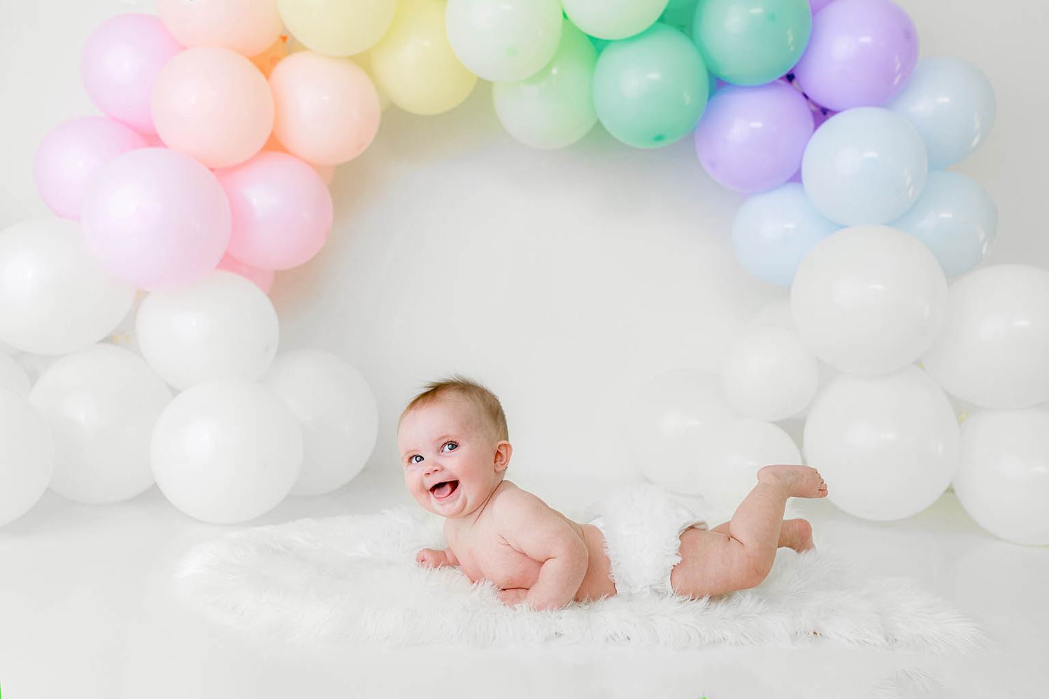 Laughing baby crawling under a vibrant pastel rainbow balloon arch on a fluffy white surface.