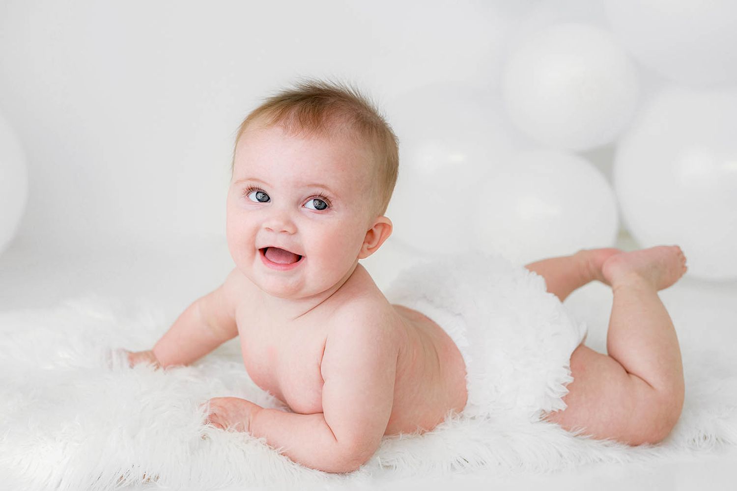 Adorable baby in diaper lying on white fluffy blanket, smiling happily surrounded by white balloons.