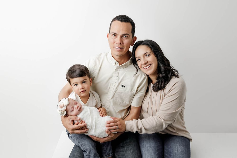 Family of four posing together in studio, parents smiling with toddler holding swaddled newborn.