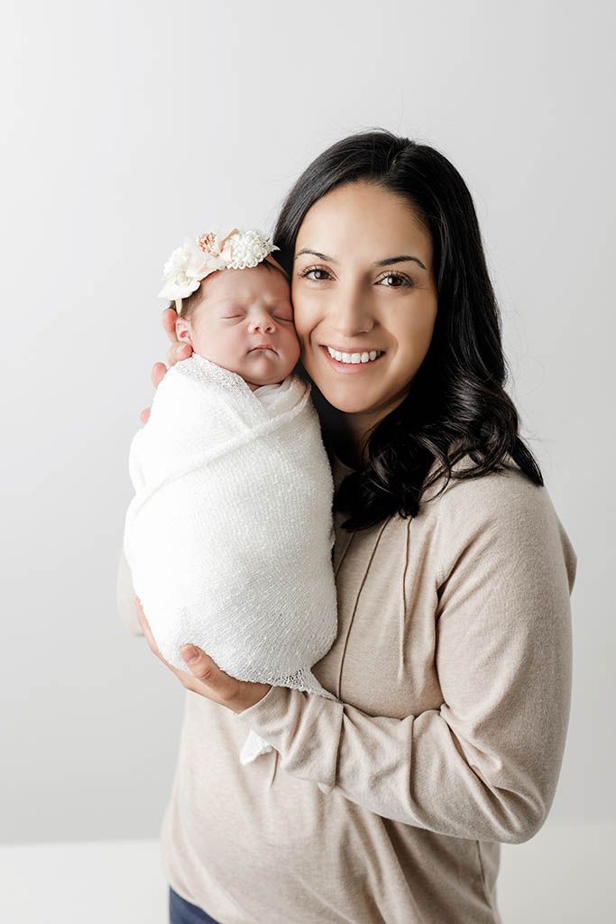 A smiling dark-haired mother holds her swaddled newborn baby girl wearing a floral headband against a white background.