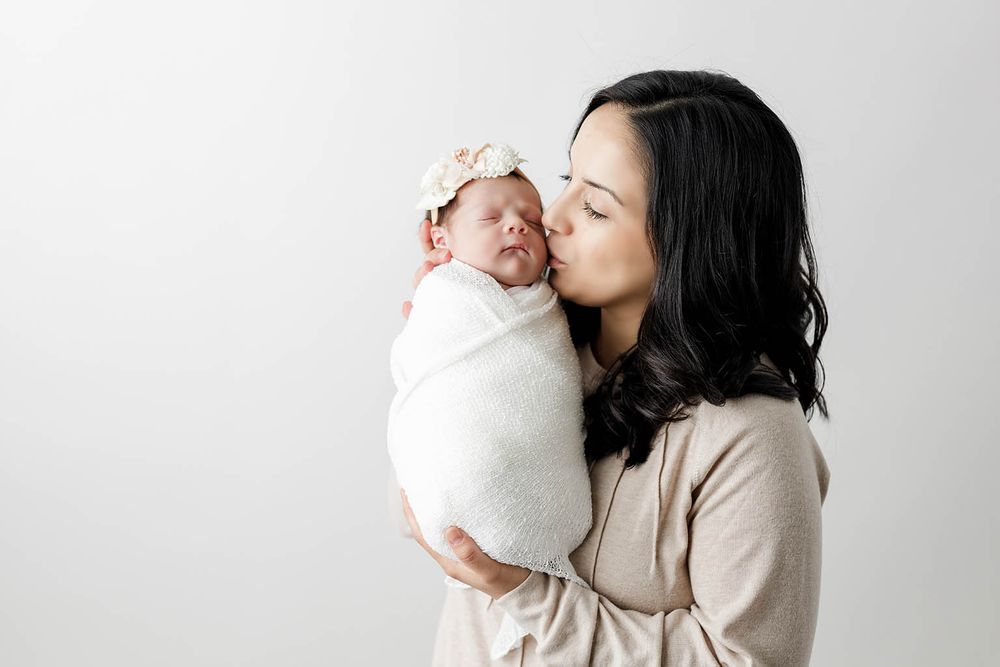 A mother tenderly kisses her sleeping newborn baby wrapped in white swaddle during a studio portrait session.