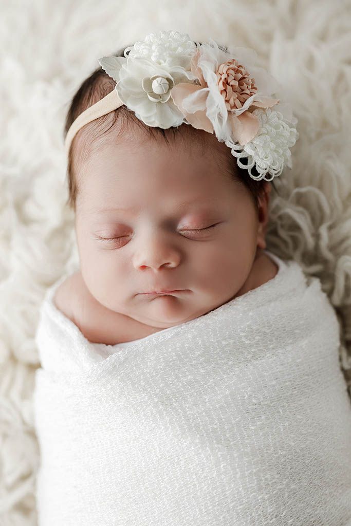 Sleeping newborn baby girl swaddled in white wrap wearing a floral headband.