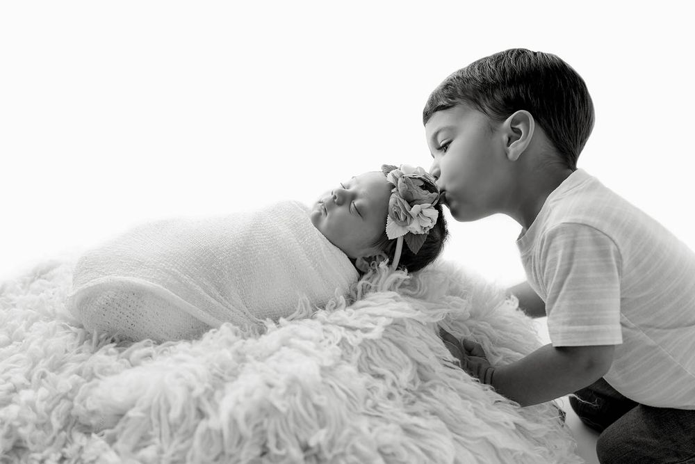A young boy lovingly kisses his sleeping newborn sibling wrapped in white during a black and white studio photo.