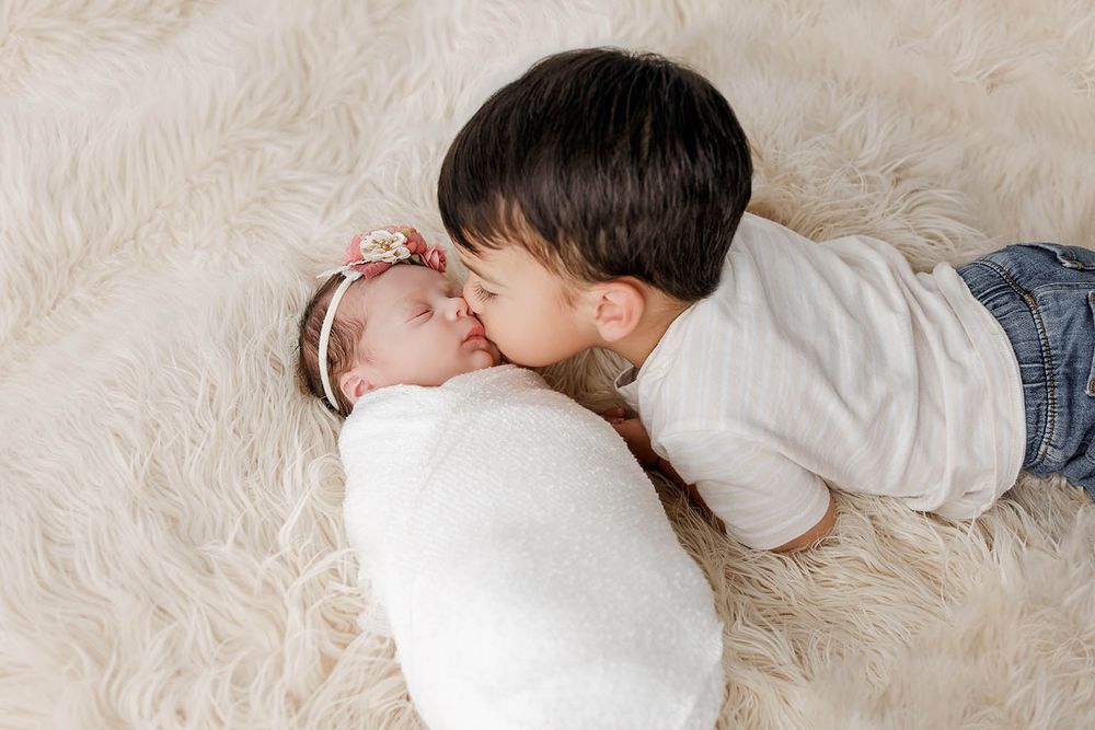 Toddler boy gently kisses newborn baby sister wrapped in white swaddle on a fluffy cream rug.