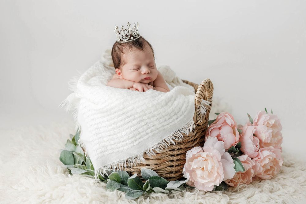 Sleeping newborn baby in a wicker basket with a tiny crown and pink peonies.